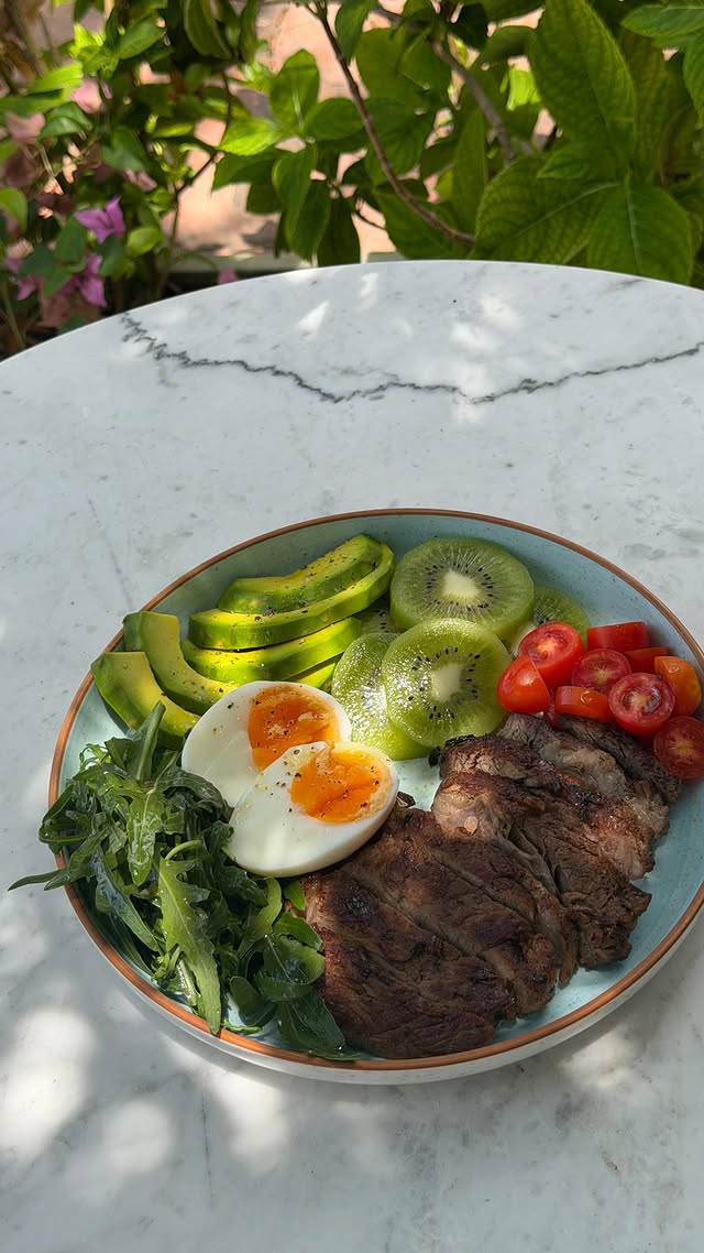 Breakfast plate on a marble table — soft-boiled egg, sliced avocado, kiwi, cherry tomatoes, grilled beef and fresh arugula in an outdoor garden setting